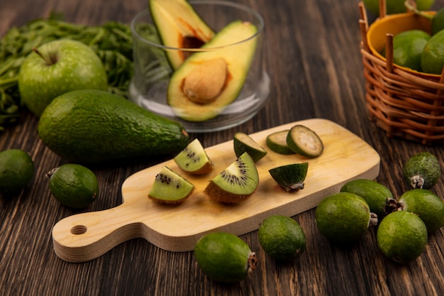 Draufsicht der frischen Scheiben der Kiwi auf einem hölzernen Küchenbrett mit halben Avocados auf einer Glasschale mit grünen Äpfeln feijoas Avocados und Petersilie lokalisiert auf einer Holzwand