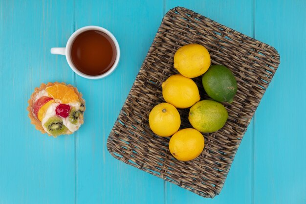 Draufsicht der frischen Zitronen auf einem Eimer mit einer Tasse Tee und Obsttorte auf einem blauen hölzernen Hintergrund
