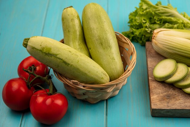 Draufsicht der frischen Zucchini auf einem Eimer mit Selleriesalat und gehackten Zucchini auf einem hölzernen Küchenbrett mit Tomaten lokalisiert auf einer blauen Holzoberfläche