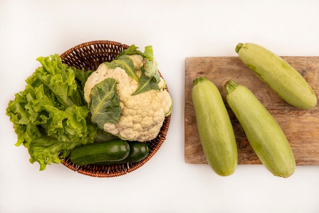 Draufsicht der frischen Zucchini lokalisiert auf einem hölzernen Küchenbrett mit Blumenkohlsalat und Gurken auf einem Eimer auf einer weißen Wand