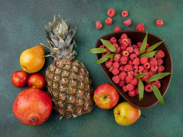 Draufsicht der Früchte als Ananas-Granatapfel-Apfel-Pfirsich-Pflaume mit Korb der Himbeere auf grüner Oberfläche