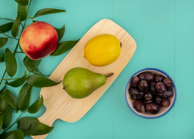 Draufsicht der Früchte als Birnenzitrone auf Schneidebrett und Schüssel Traubenbeeren und Pfirsich mit Blättern auf blauem Hintergrund