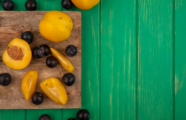 Draufsicht der Früchte als geschnittene und ganze Aprikosen und Schlehenbeeren auf Schneidebrett und auf grünem Hintergrund mit Kopienraum