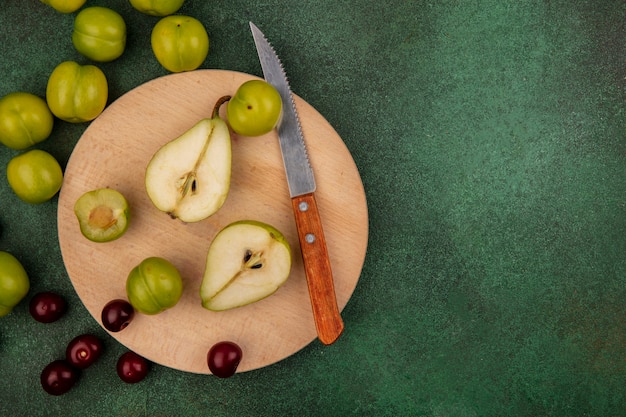 Kostenloses Foto draufsicht der früchte als halb geschnittene birne und pflaume mit messer auf schneidebrett und kirschen auf grünem hintergrund mit kopienraum