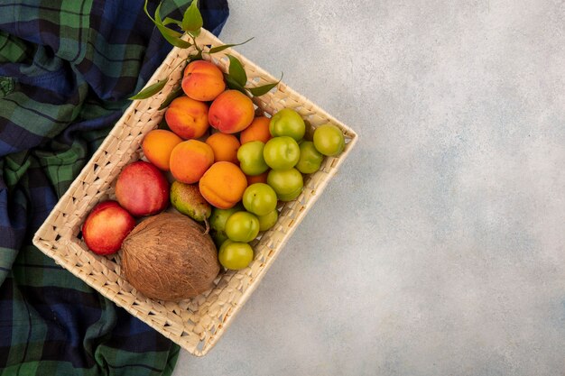 Draufsicht der Früchte als Pfirsich-Aprikosen-Pflaumen-Kokosnuss im Korb auf kariertem Stoff und weißem Hintergrund mit Kopienraum