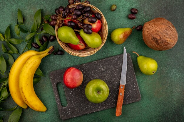 Draufsicht der Früchte als Pfirsichapfel mit Messer auf Schneidebrett und Birnen-Kokosnuss-Traubenbanane mit Blättern auf grünem Hintergrund