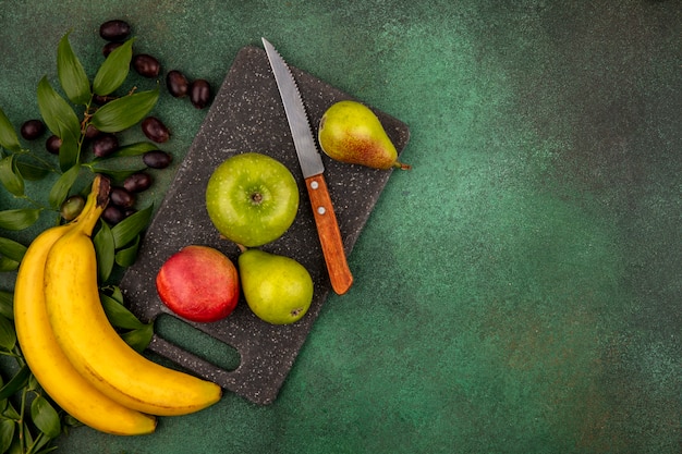 Draufsicht der Früchte als Pfirsichapfelbirne mit Messer auf Schneidebrett und Traubenbanane mit Blättern auf grünem Hintergrund mit Kopienraum