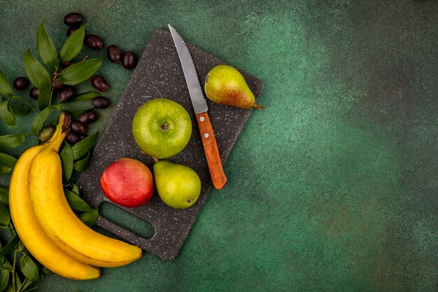 Draufsicht der Früchte als Pfirsichapfelbirne mit Messer auf Schneidebrett und Traubenbanane mit Blättern auf grünem Hintergrund mit Kopienraum