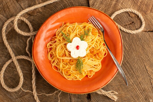 Draufsicht der gekochten italienischen Nudeln mit Grüns innerhalb des orangefarbenen Tellers auf dem hölzernen Schreibtisch