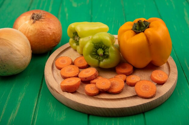 Draufsicht der gelben und grünen Paprika auf einem hölzernen Küchenbrett mit gehackten Karotten mit Zwiebeln, die auf einer grünen Holzwand lokalisiert werden