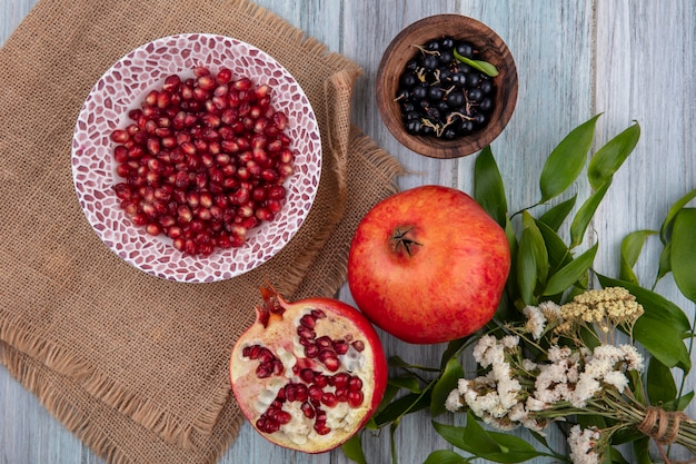 Draufsicht der Granatapfelbeeren in der Schüssel auf Sackleinen mit ganzen und halben und Schüssel Brombeere mit Blumen auf Holzoberfläche