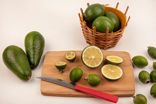Draufsicht der grünen Kalkscheiben auf einem hölzernen Küchenbrett mit Messer mit Limetten auf einem Eimer mit Feijoas und Avocados lokalisiert auf einer weißen Wand