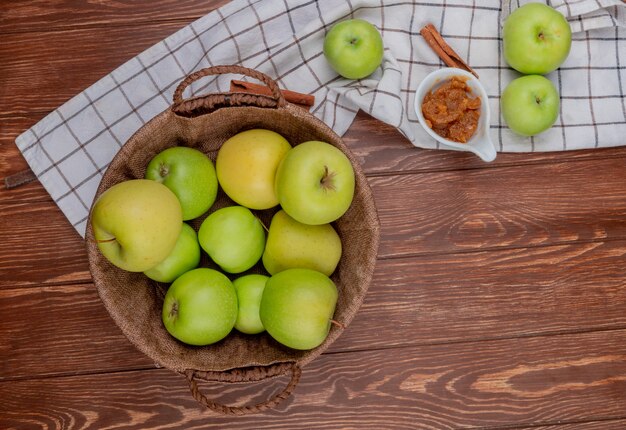 Draufsicht der grünen und gelben Äpfel im Korb mit Apfelmarmelade und Zimt auf kariertem Stoff und hölzernem Hintergrund