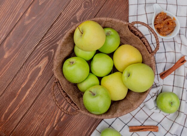 Draufsicht der grünen und gelben Äpfel im Korb mit Apfelmarmelade und Zimt auf kariertem Stoff und Holztisch mit Kopienraum