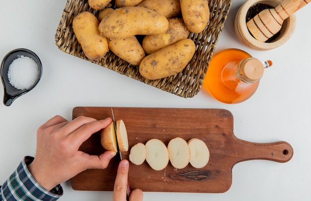 Draufsicht der Hände, die Kartoffel mit Messer auf Schneidebrett und anderen in Platte Butter Salz schwarzen Pfeffer auf weißer Oberfläche schneiden