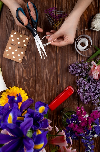 Draufsicht der Hände mit der Schere, die eine Seilpostkarten-Büroklammer und einen Blumenstrauß der lila Irisblumen auf hölzernem Hintergrund schneidet