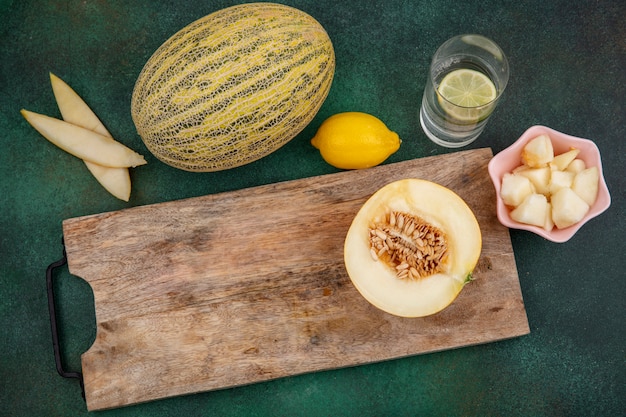 Kostenloses Foto draufsicht der halbierten melone auf einem hölzernen küchenbrett mit melonenscheiben auf einer rosa schüssel auf grüner oberfläche