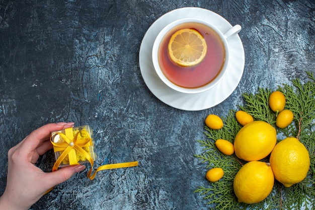 Draufsicht der Hand, die ein Geschenk und eine Tasse schwarzen Tee mit Zitrone neben der Zitrusfruchtsammlung auf Tannenzweigen auf dunklem Hintergrund hält