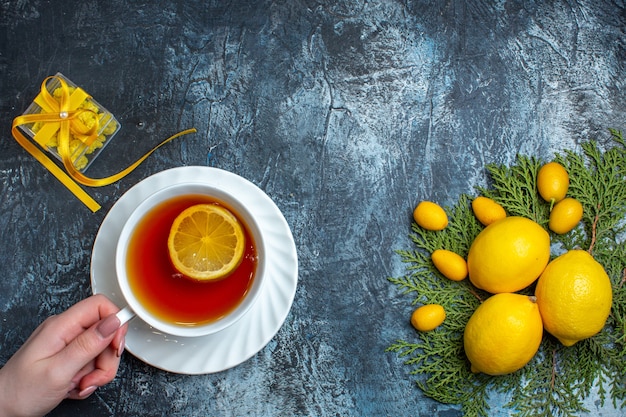 Draufsicht der Hand, die eine Tasse schwarzen Tee mit Zitrone und gelber Geschenkbox neben der Zitrusfruchtsammlung auf Tannenzweigen auf dunklem Hintergrund hält