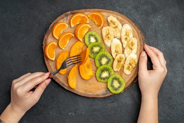 Kostenloses Foto draufsicht der hand, die natürliches organisches frisches obst mit gabel auf schneidebrett auf dunklem hintergrund nimmt