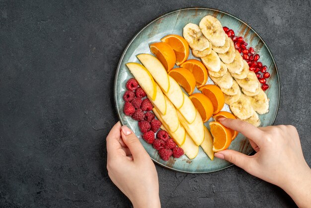 Draufsicht der Hand, die Orangenscheiben-Sammlung von gehackten frischen Früchten auf einem blauen Teller auf schwarzem Tisch nimmt