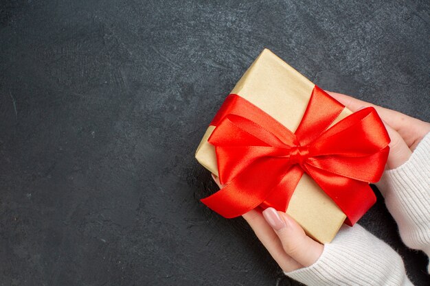 Draufsicht der Hand, die schönes Geschenk mit bogenförmigem rotem Band auf der Seite auf dunklem Hintergrund hält