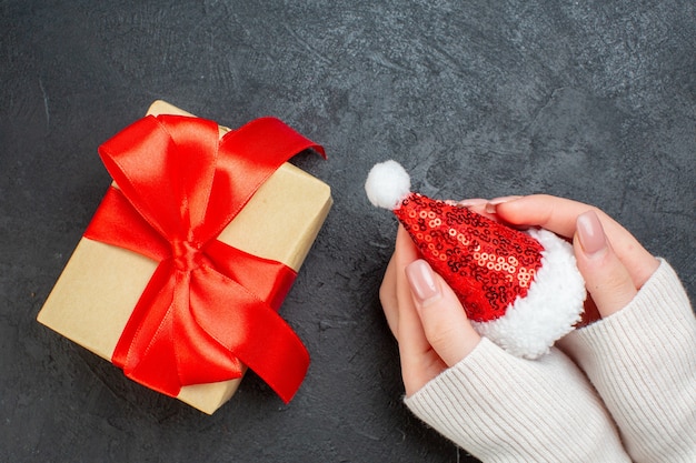 Draufsicht der Hand, die Weihnachtsmannhut und schönes Geschenk mit bogenförmigem Band auf dunklem Hintergrund hält