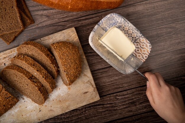 Draufsicht der Handhandschneidebutter der Frau mit Messer und geschnittenem Sandwichbrot auf Schneidebrett auf hölzernem Hintergrund