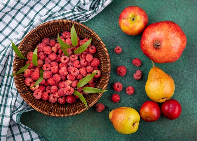 Draufsicht der Himbeeren im Korb auf kariertem Stoff und Muster der Granatapfelpfirsichapfelpflaume auf grüner Oberfläche