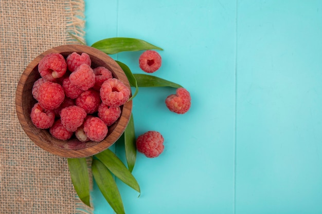 Draufsicht der Himbeeren in der Schüssel auf Sackleinen mit Blättern auf blauer Oberfläche