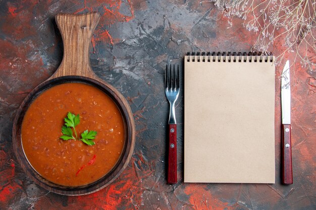 Draufsicht der klassischen Tomatensuppe auf einem braunen Schneidebrett und Notizbuch auf gemischtem Farbhintergrund