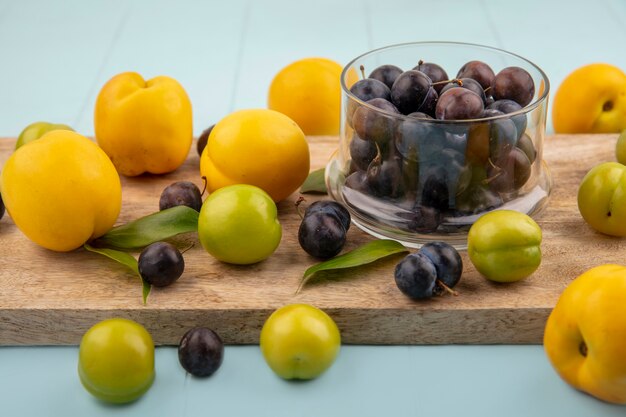 Draufsicht der kleinen sauren blau-schwarzen Fruchtschleifen auf einer Glasschale auf einem hölzernen Küchenbrett mit grünen Kirschpflaumen mit gelben Pfirsichen auf einem blauen Hintergrund