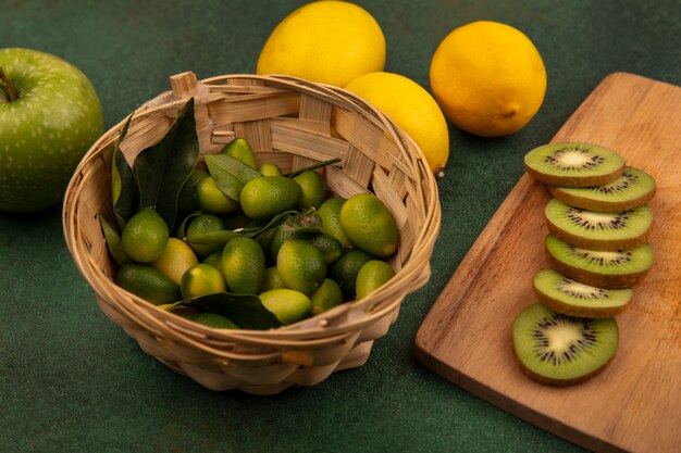 Draufsicht der köstlichen Kiwischeiben auf einem hölzernen Küchenbrett mit Kinkans auf einem Eimer mit Zitronen und Apfel lokalisiert auf einer grünen Oberfläche