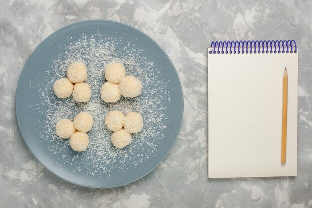 Draufsicht der köstlichen Kokosnussbonbons innerhalb der blauen Platte mit Notizblock auf weißer Oberfläche