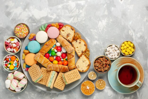 Draufsicht der köstlichen Süßekompositionskuchenscheiben Macarons-Bonbons mit Tasse Tee auf weißer Oberfläche