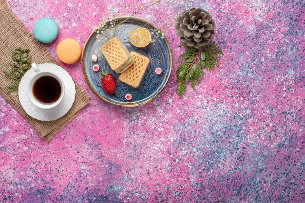 Draufsicht der köstlichen Waffeln mit Tasse Tee und Macarons auf rosa Oberfläche