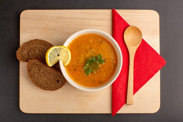 Draufsicht der leckeren Gemüsesuppe innerhalb Platte mit Zitronenscheibe und Brotlaib auf der dunklen Oberfläche