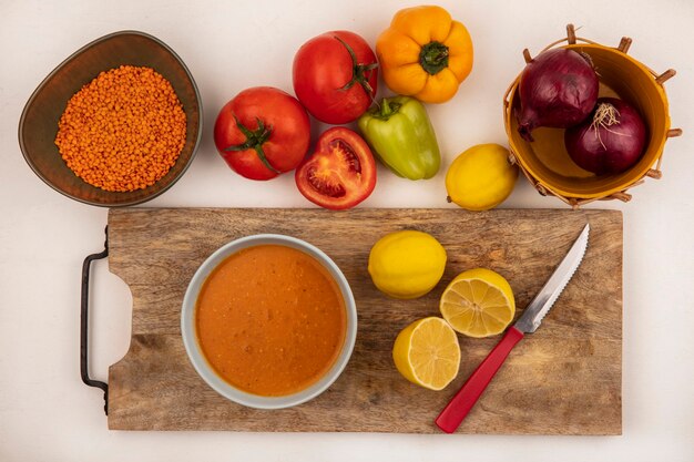 Draufsicht der leckeren Linsensuppe auf einer Schüssel auf einem hölzernen Küchenbrett mit Zitronen mit Messer mit roten Zwiebeln auf einem Eimer mit Tomaten und Paprika lokalisiert auf einer weißen Wand