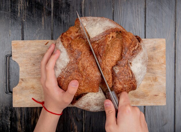 Draufsicht der männlichen Hände, die Cob Brot mit Messer auf Schneidebrett auf hölzernem Hintergrund schneiden