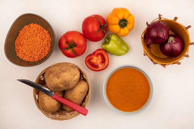 Draufsicht der nahrhaften Linsensuppe auf einer Schüssel mit roten Zwiebeln auf einem Eimer mit Kartoffeln auf einem Eimer mit Messer mit Paprika und Tomaten lokalisiert auf einer weißen Wand