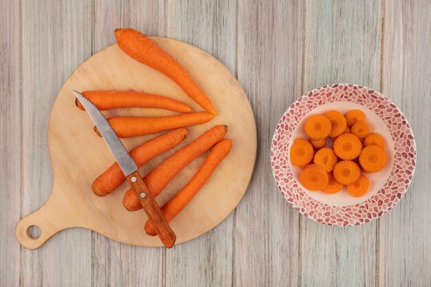 Draufsicht der orange Karotten auf einem hölzernen Küchenbrett mit Messer mit gehackten Karotten auf einer Schüssel auf einem grauen hölzernen Hintergrund