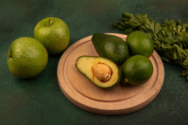 Draufsicht der organischen Avocados auf einem hölzernen Küchenbrett mit Limetten mit grünen Äpfeln und Petersilie lokalisiert auf einem grünen Hintergrund