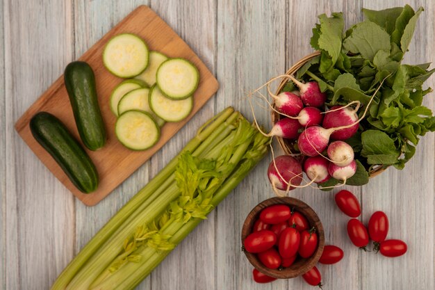 Draufsicht der organischen Tomaten auf einer hölzernen Schüssel mit Radieschen auf einem Eimer mit Gurken auf einem hölzernen Küchenbrett mit Tomaten und Sellerie lokalisiert auf einer grauen Holzoberfläche