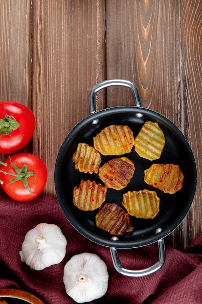 Draufsicht der Pfanne mit Kartoffelchips und -gemüse als Knoblauch und Tomate auf hölzernem Hintergrund mit Kopienraum