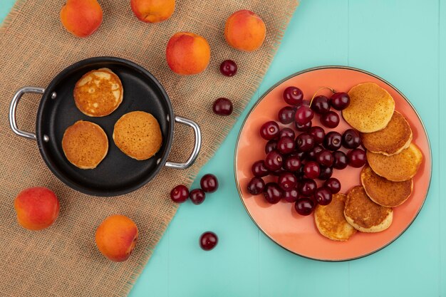 Draufsicht der Pfannkuchen in der Pfanne und im Teller mit Kirschen und Aprikosen auf Sackleinen und auf blauem Hintergrund