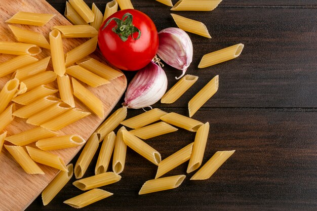 Draufsicht der rohen Nudeln auf einem Schneidebrett mit Tomate und Knoblauch auf einer Holzoberfläche