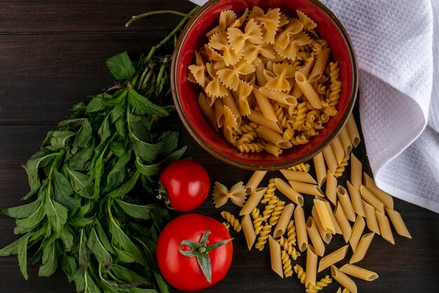 Draufsicht der rohen Nudeln in einer Schüssel mit Tomaten und einem Bündel Minze auf einer Holzoberfläche