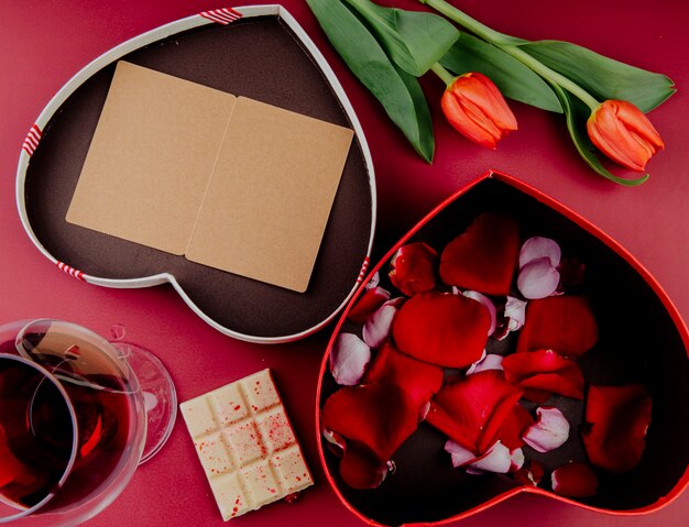 Draufsicht der roten Farbe Tulpenblumen mit herzförmiger Geschenkbox mit einer offenen Postkarte und einer Box gefüllt mit Rosenblättern und weißer Schokolade mit einem Glas Wein auf rotem Hintergrund