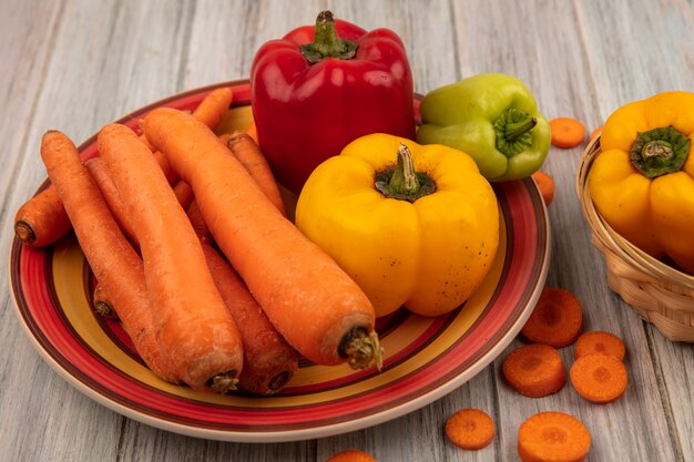 Draufsicht der roten gelben und grünen Paprika auf einem Teller mit Karotten mit gelben Paprika auf einem Eimer auf einer grauen Holzoberfläche