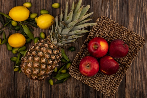 Draufsicht der roten Äpfel auf einem Weidentablett mit Ananas-Zitronen und Kinkans lokalisiert auf einer Holzoberfläche